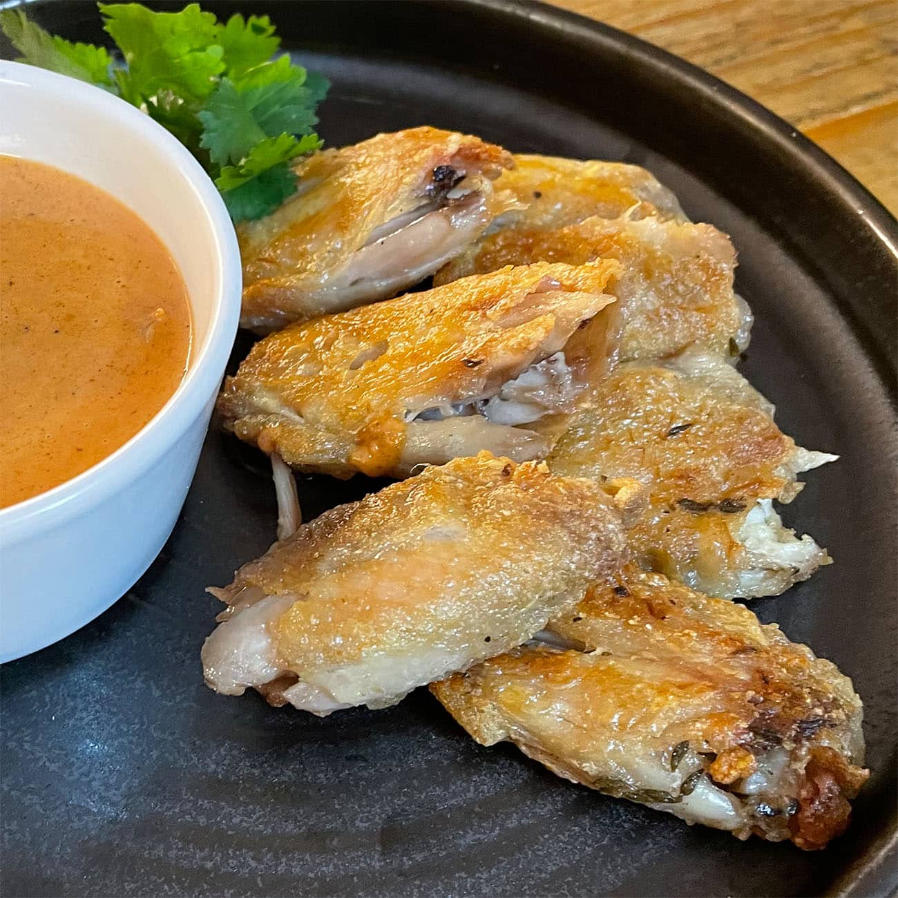 Crispy baked chicken wings arranged on a black plate, served with a pot of orange dipping sauce and a garnish of fresh coriander.