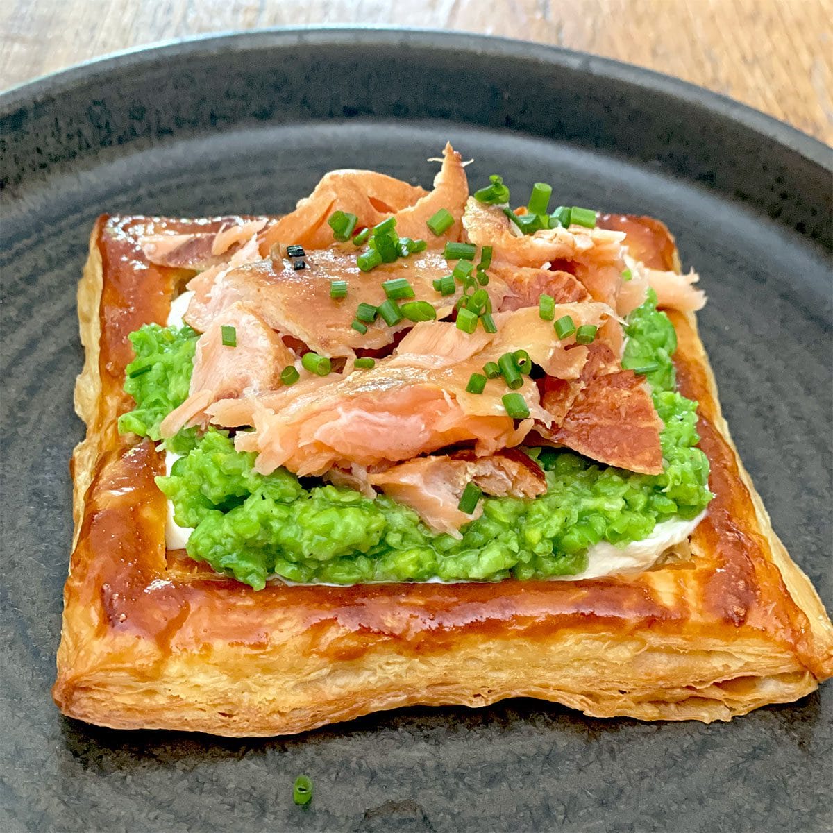 A square puff pastry topped with a layer of mashed peas, flaked smoked salmon, and chopped chives, served on a dark round plate.