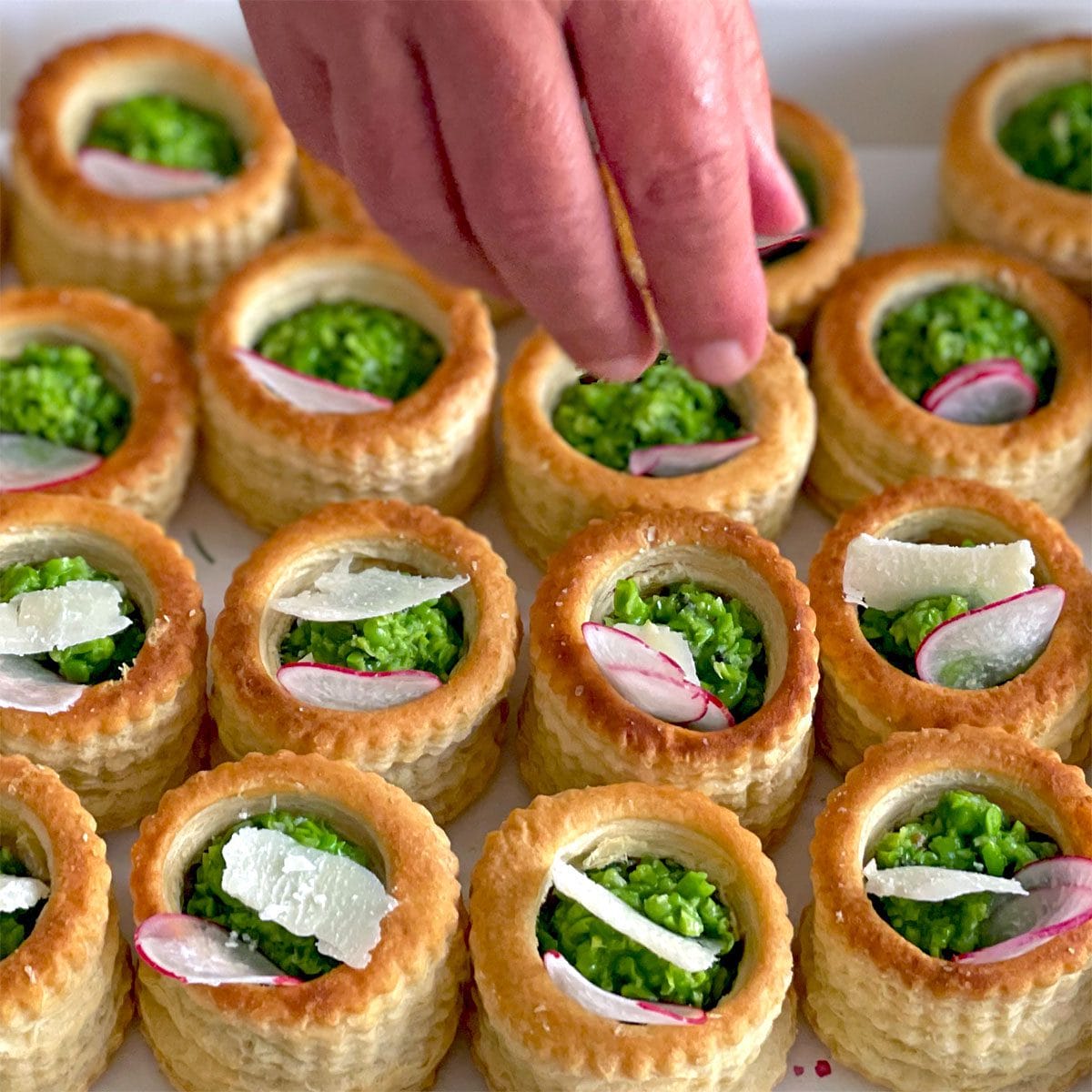 A hand garnishes round puff pastry shells filled with garden peas, topped with radish slices and cheese shavings, arranged neatly on a tray.