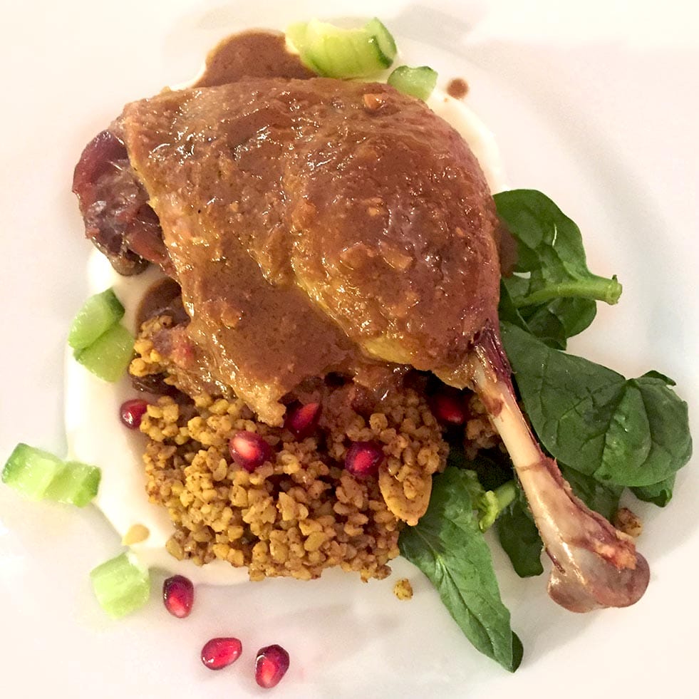 A cooked duck leg with brown sauce is served on a bed of spinach, bulgur or couscous, pomegranate seeds, and diced celery, plated on a white plate.