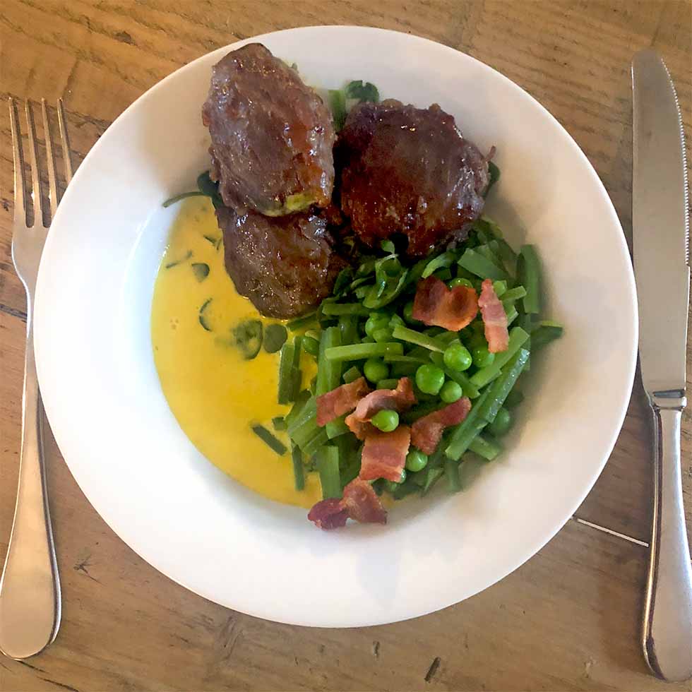 A white plate with three pieces of glazed meat, green beans and peas topped with bacon pieces, and yellow sauce, set on a wooden table with a fork and knife beside the plate.