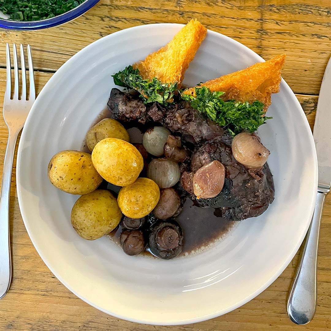 A plate with boiled baby potatoes, pearl onions, mushrooms, braised meat in dark sauce, and two triangular pieces of toast topped with chopped herbs. Knife and fork are placed on either side of the plate.