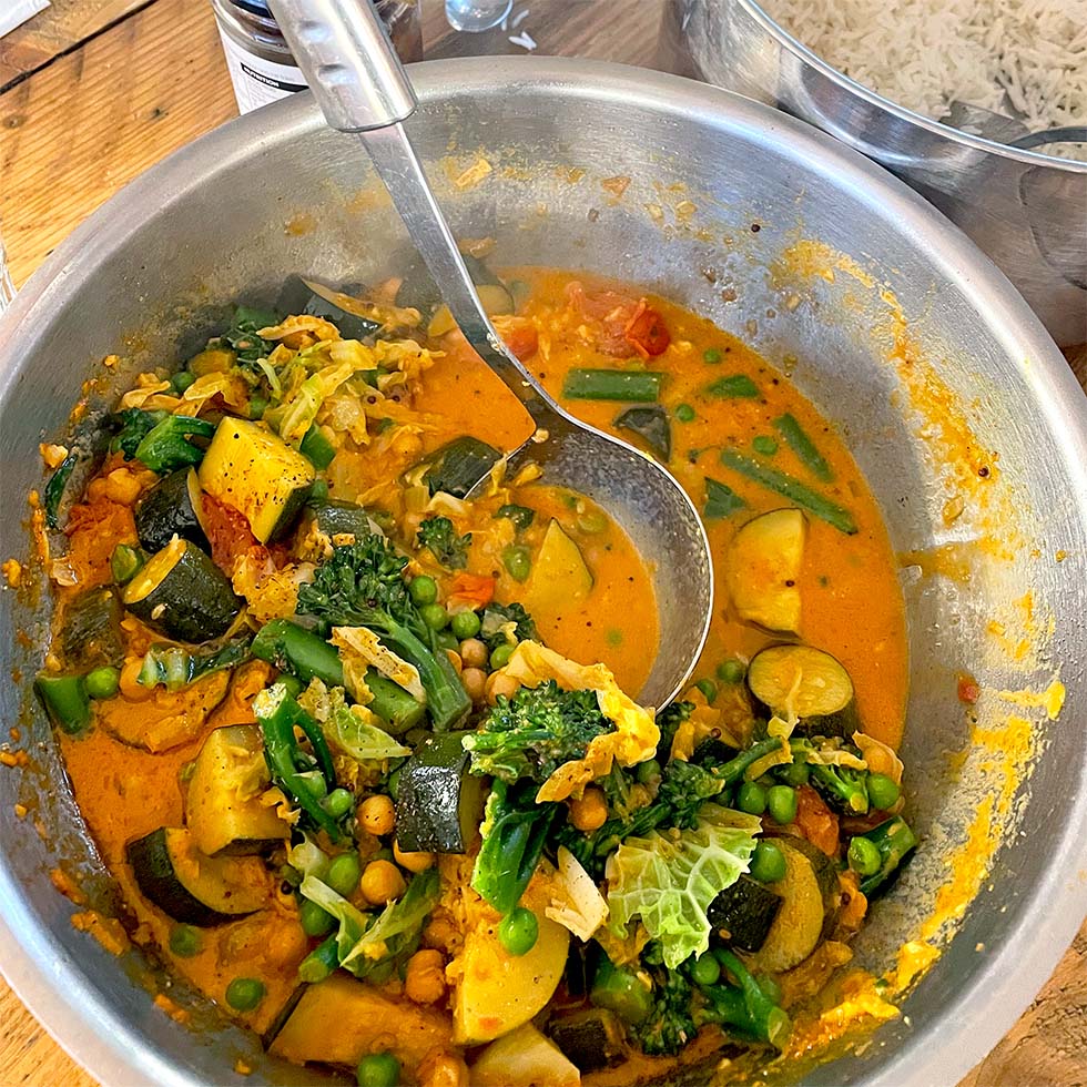 A large metal bowl filled with vibrant vegetable curry, including courgette, broccoli, peas, green beans, and chickpeas in a rich orange sauce, with a ladle resting inside. A bowl of white rice is in the background.