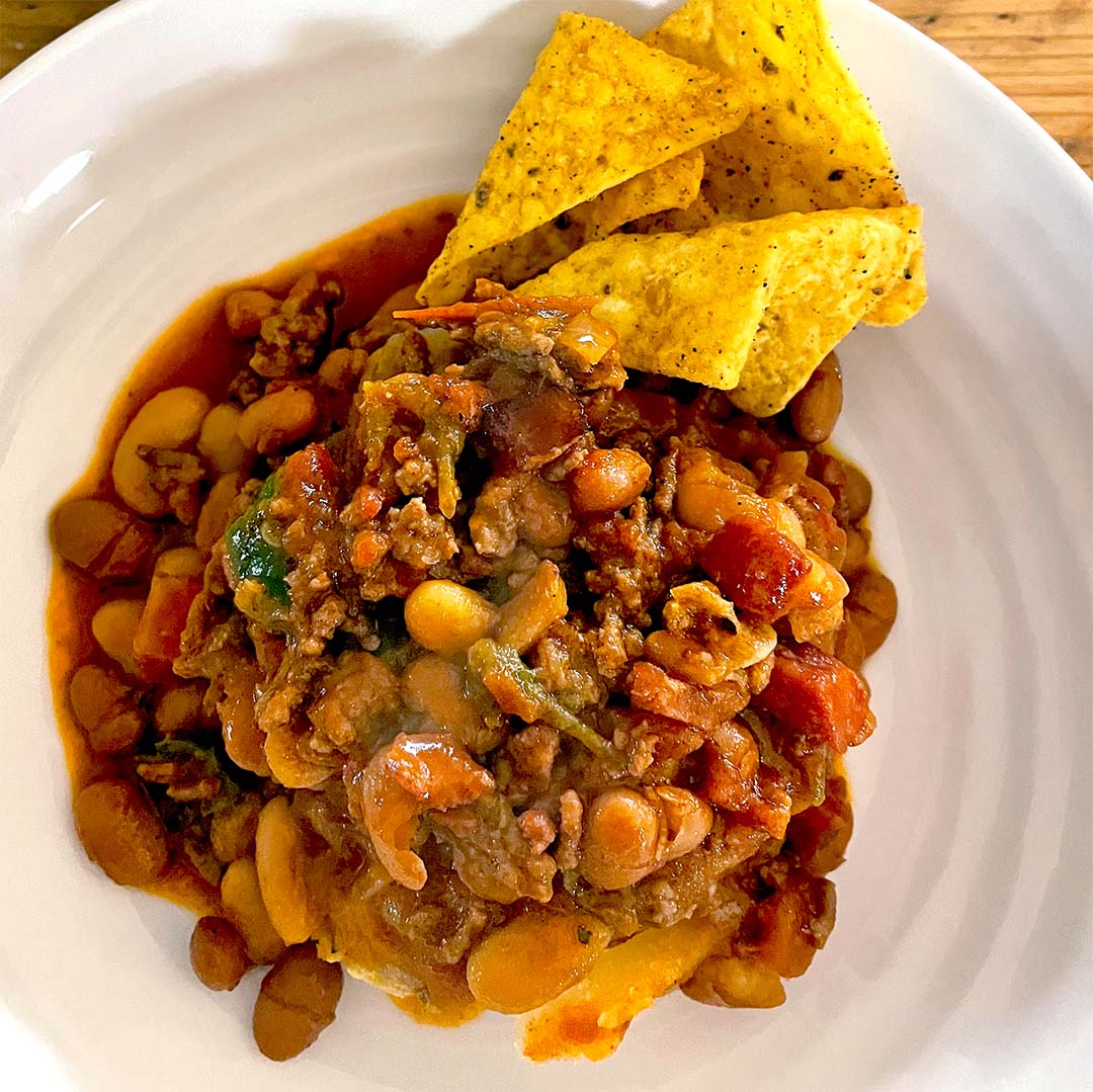 A plate of chilli with beans, minced meat, tomatoes, and vegetables, topped with three yellow corn tortilla crisps on the side. The chilli appears hearty and saucy, served on a white plate.