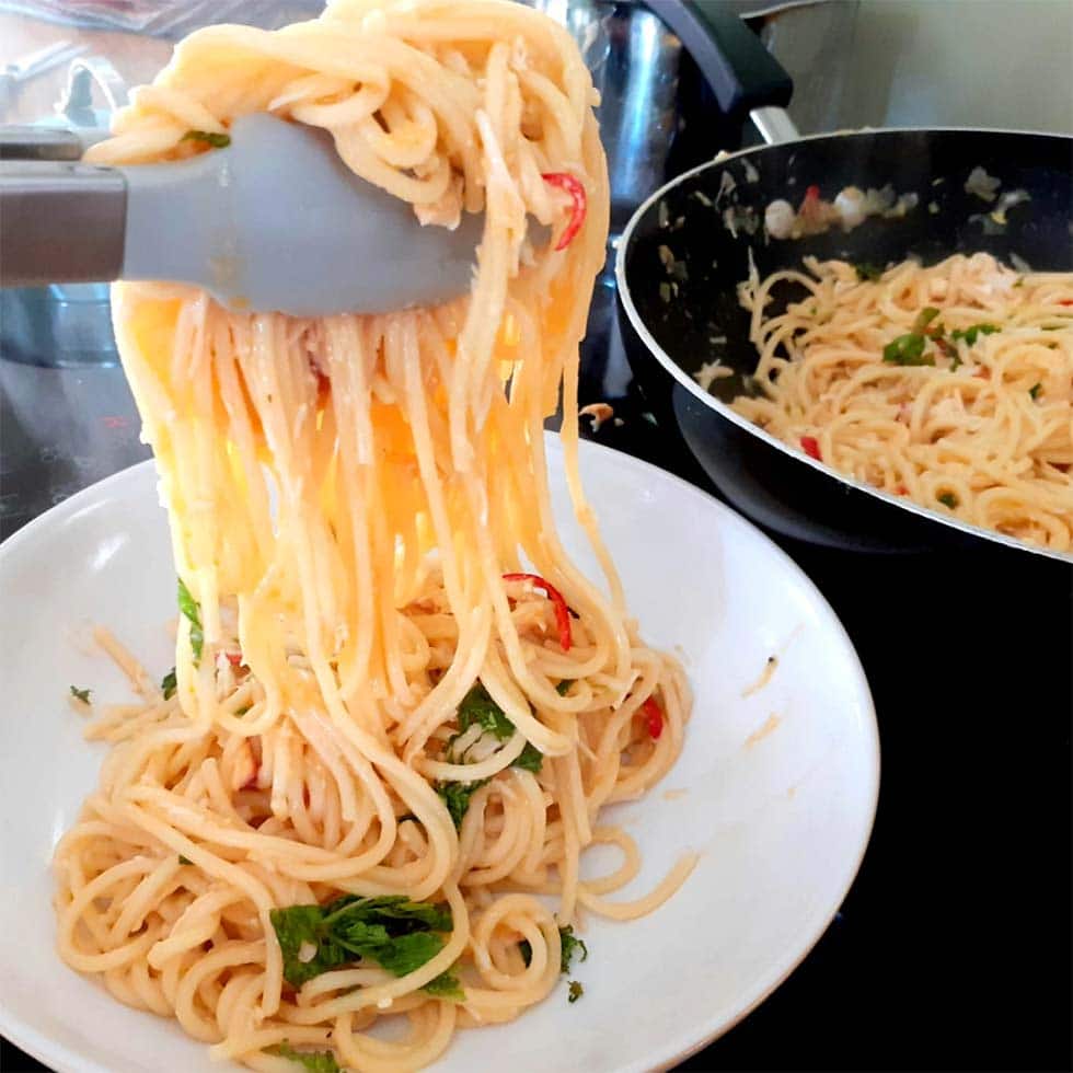 A close-up of spaghetti being served from a frying pan into a white bowl using kitchen tongs. The pasta is mixed with herbs and sliced red chilli peppers.