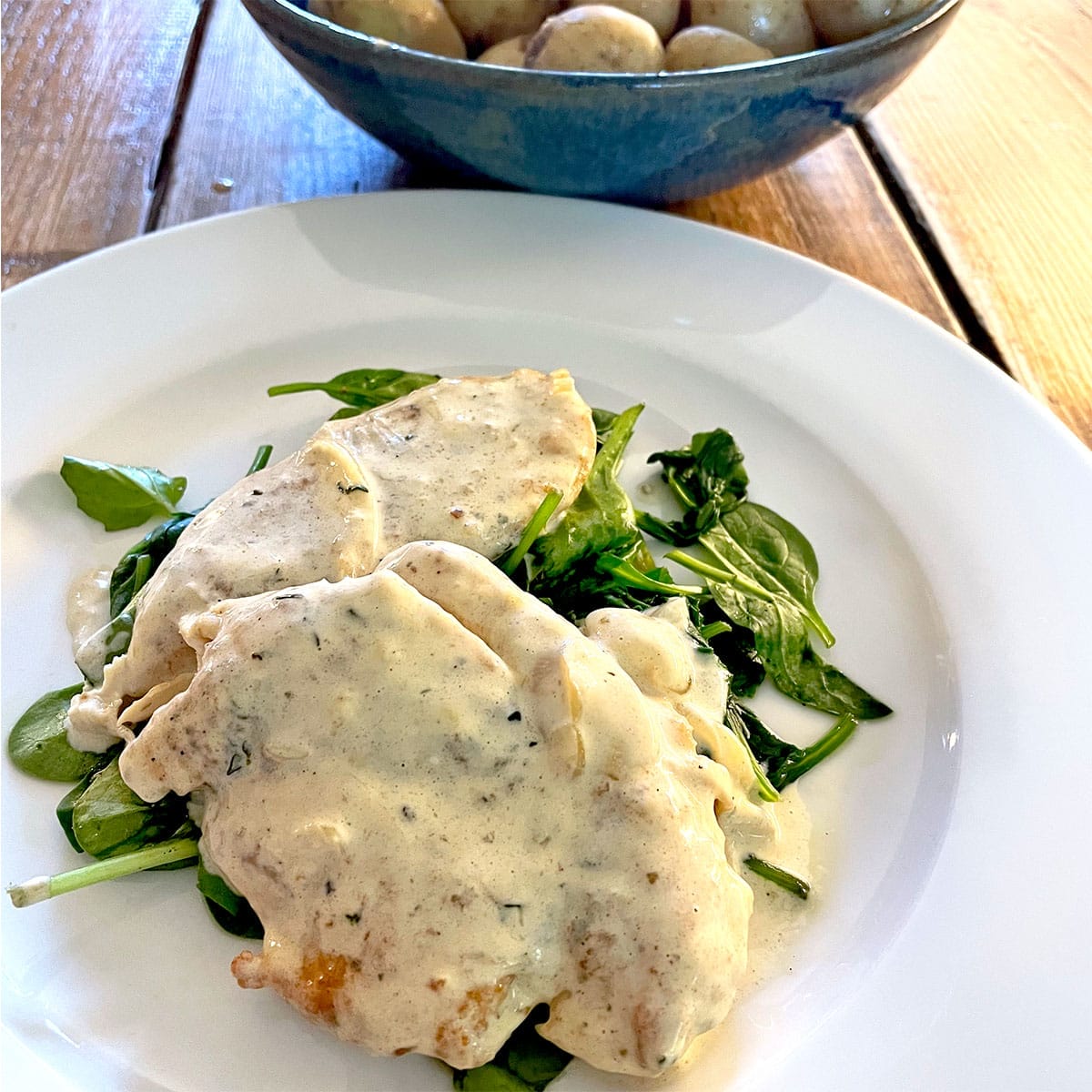 Two pieces of chicken breast in a creamy white herb sauce served on a bed of fresh spinach leaves on a white plate, with a bowl of whole baby potatoes in the background.