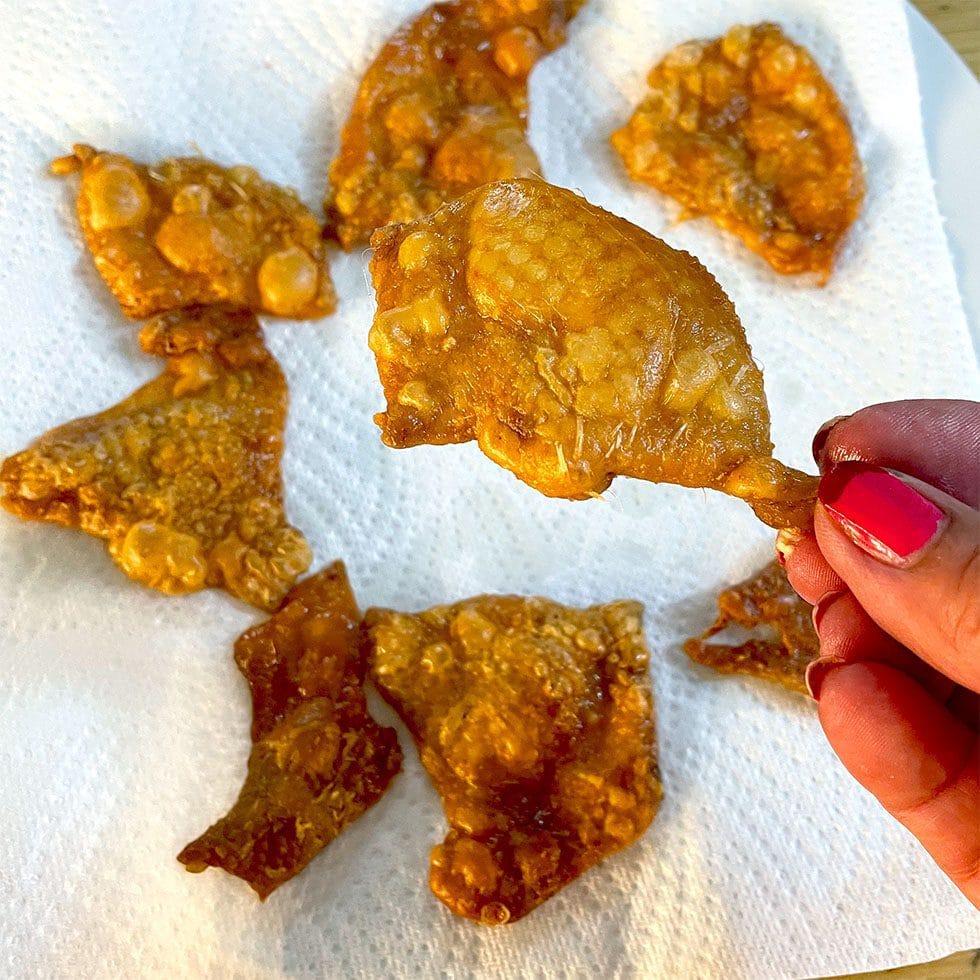 A hand with pink-painted nails holds a crispy, golden-brown fried chicken skin over a kitchen roll covered with more pieces of fried chicken skin.