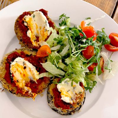 Three breaded aubergine slices topped with tomato sauce and melted cheese are served on a white plate with a side salad of mixed leaves and cherry tomatoes.