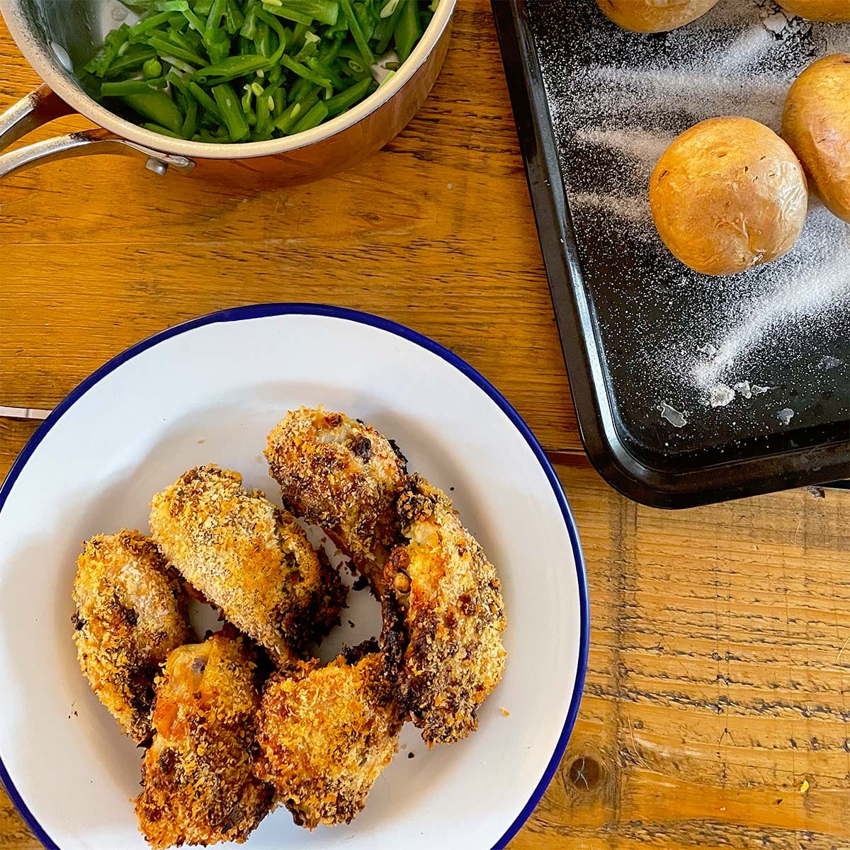 A plate of crispy breaded chicken pieces, a saucepan of green beans, and a baking tray with baked potatoes on a wooden table.