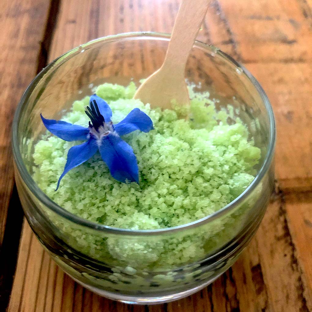 A glass beaker filled with light green granulated pudding, topped with a vibrant blue edible flower, and a small wooden spoon resting inside. The beaker sits on a rustic wooden table.
