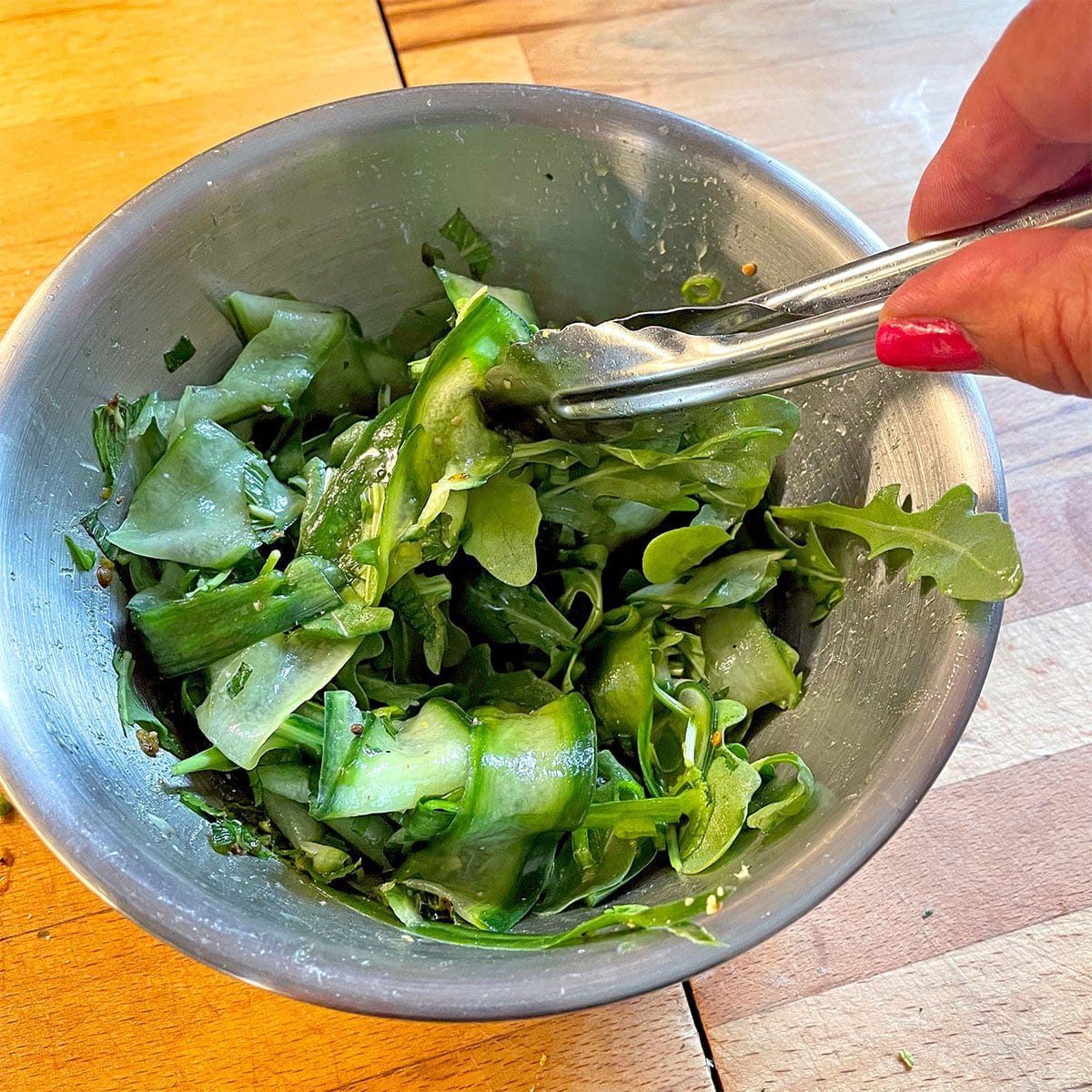 A hand with red nail varnish holds tongs, tossing a green salad with cucumber ribbons and leafy greens in a metal bowl on a wooden surface.