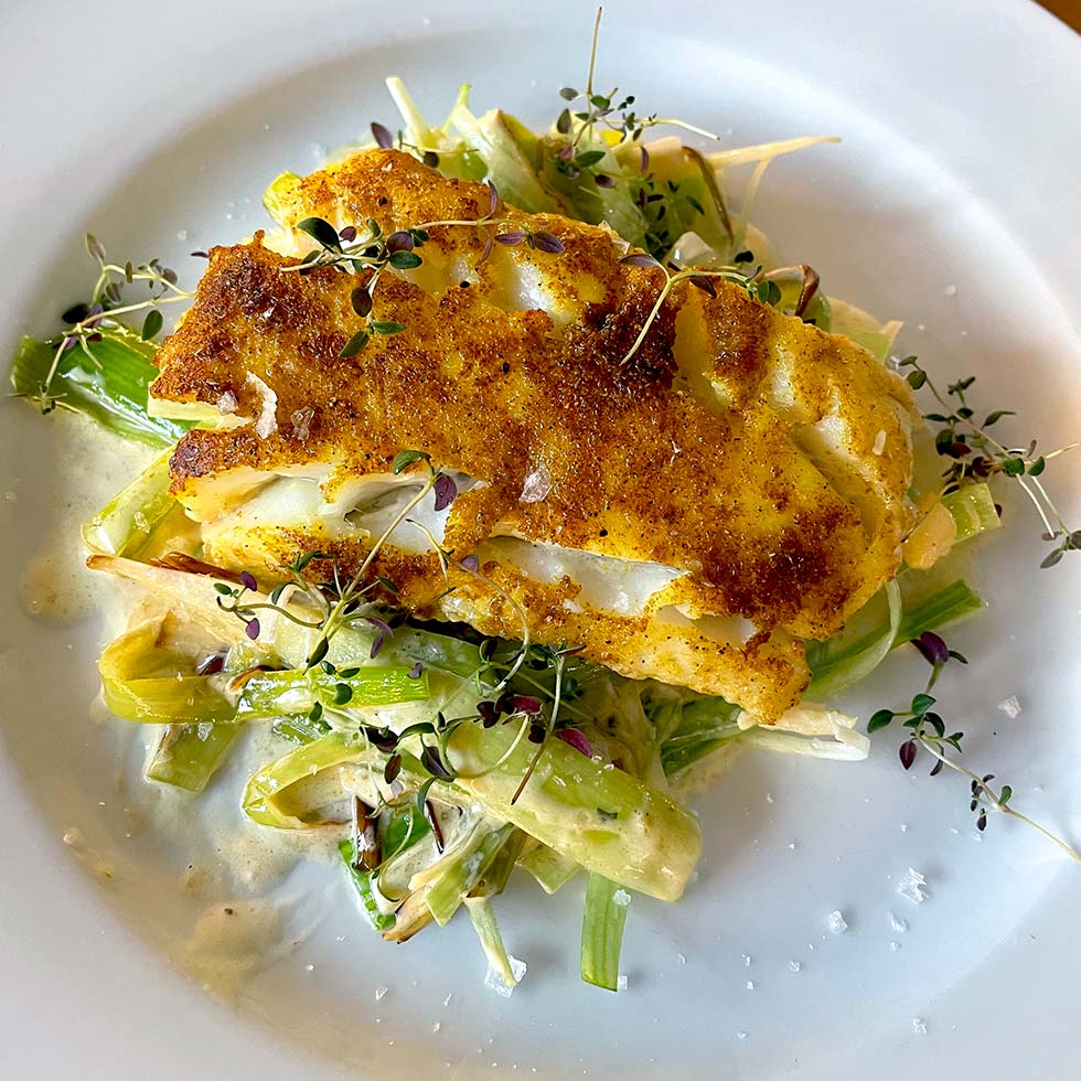 A golden-brown, pan-seared fish fillet sits atop a creamy medley of sautéed vegetables, garnished with fresh microgreens, on a white plate.