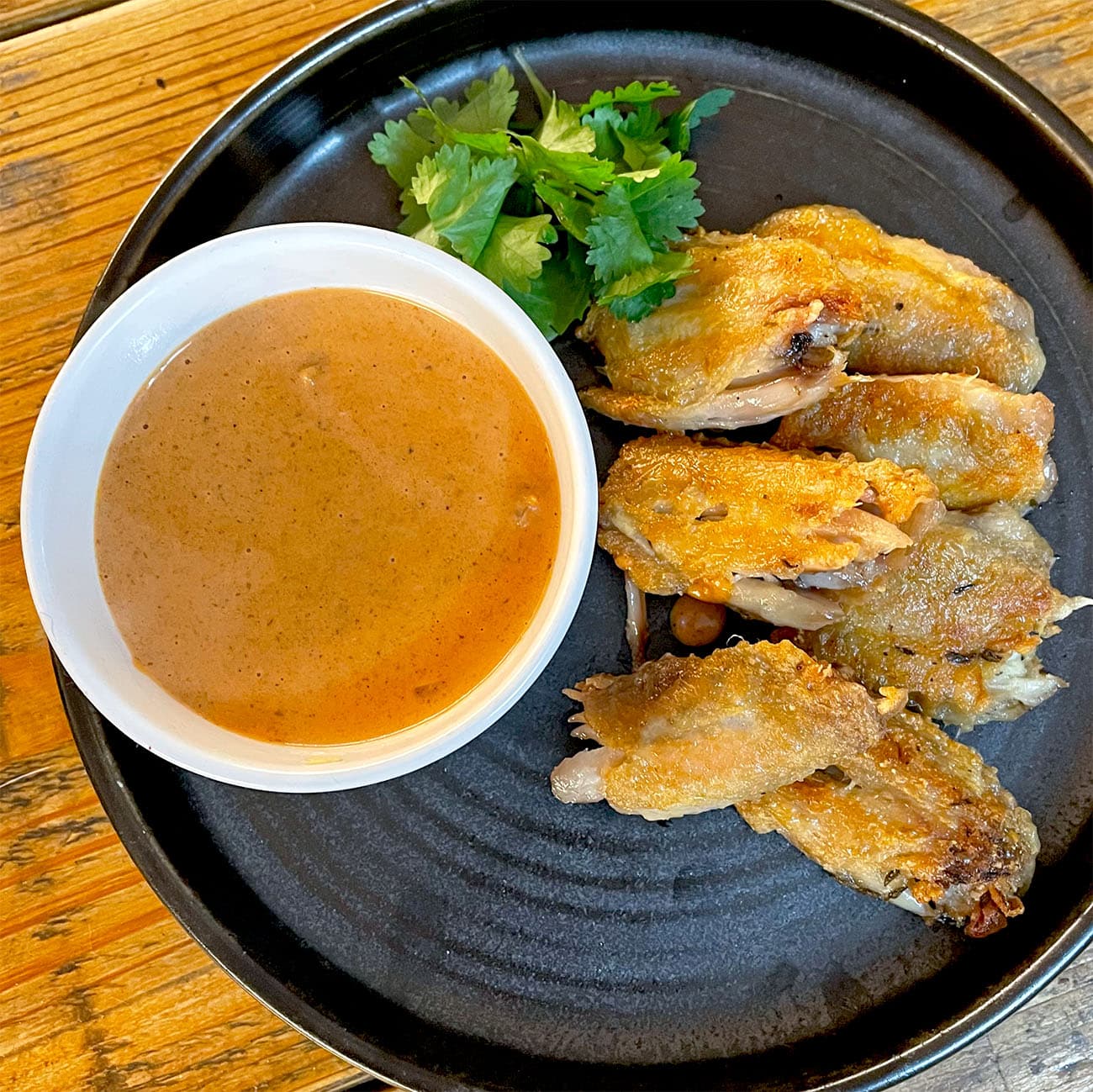 A black plate with crispy chicken wings, a garnish of coriander, and a white bowl filled with brown dipping sauce, set on a wooden table.