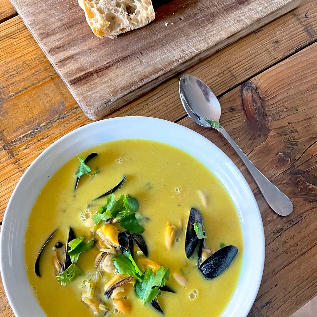 A white bowl of yellow seafood soup garnished with coriander sits on a wooden table next to a metal spoon and a chopping board with a piece of crusty bread. Black mussel shells are visible in the soup.