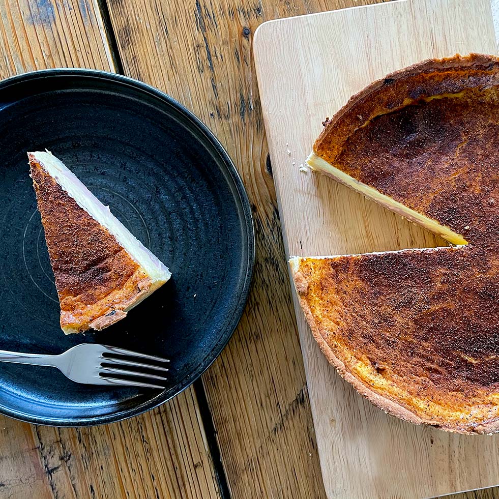 A slice of custard tart on a black plate with a fork beside it sits next to the rest of the tart, which is on a wooden chopping board with one slice missing. The surface of the tart appears caramelised.