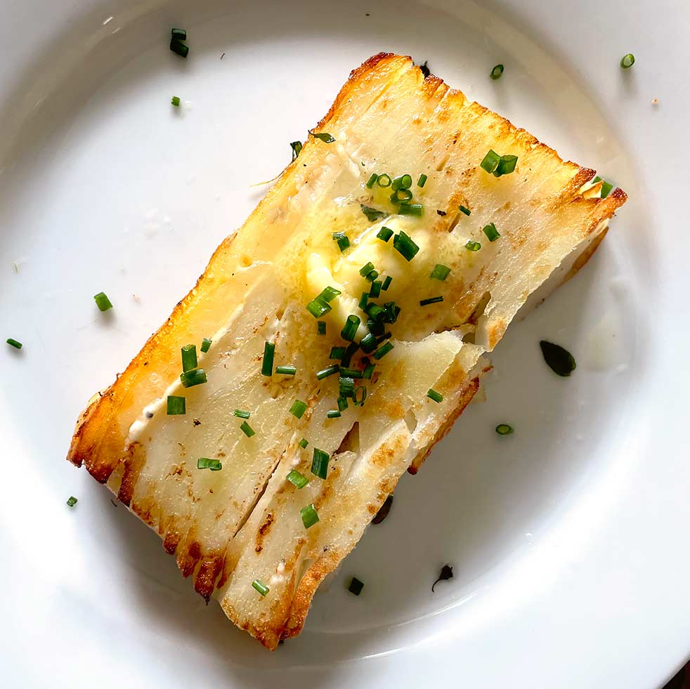 A rectangular slice of layered, golden-brown potato garnished with chopped chives and a pat of melted butter, served on a white plate.