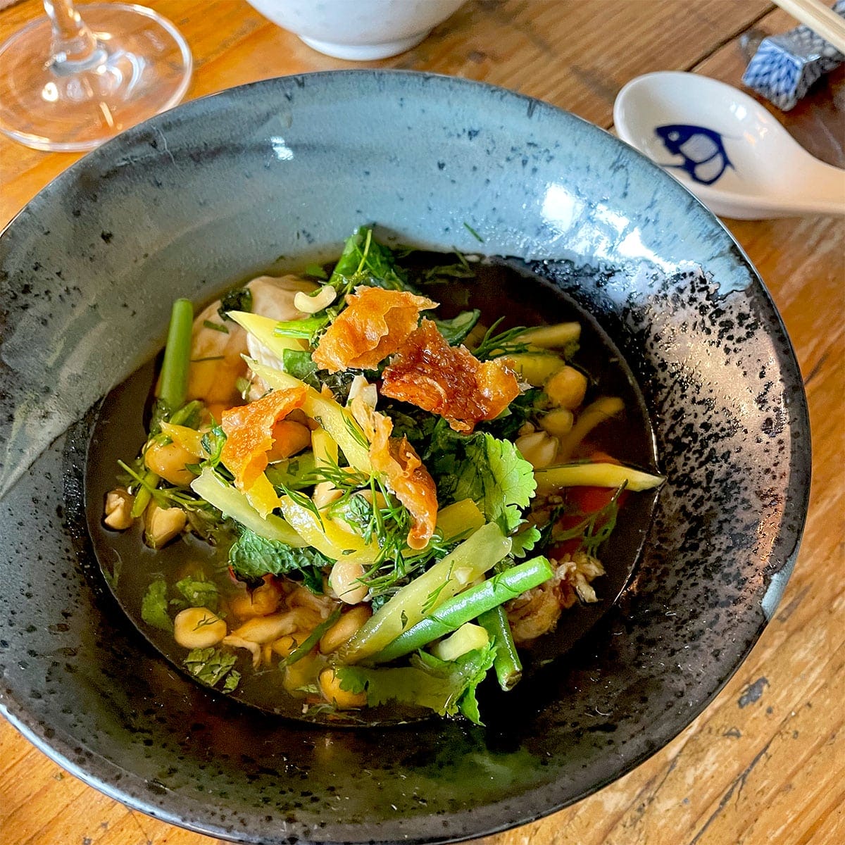 A bowl of vegetable soup garnished with fresh herbs and crispy toppings sits on a wooden table, with a decorative spoon and glass visible nearby.