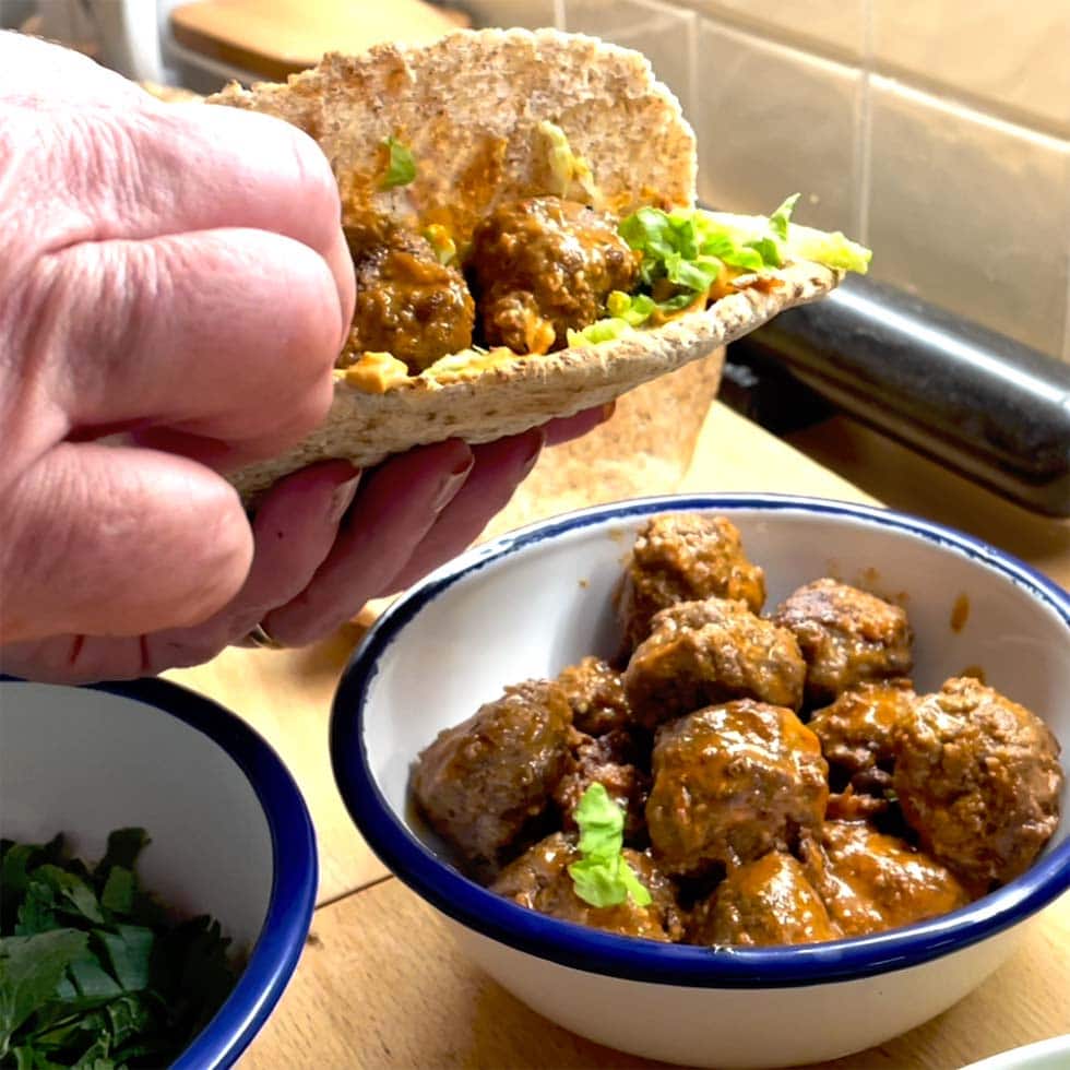 A hand holds a pitta filled with meatballs, lettuce, and sauce. Below, a bowl contains more saucy meatballs, and a small bowl of chopped herbs is nearby on a wooden worktop.
