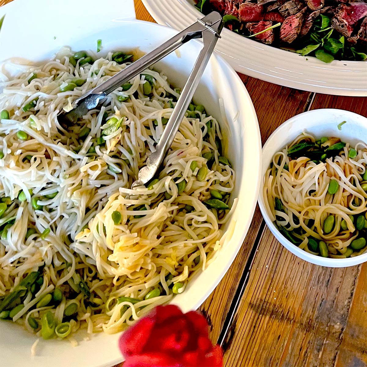 A large white bowl of noodle salad with green vegetables and metal tongs sits on a wooden table next to a smaller bowl of salad and a plate of sliced beef over greens.