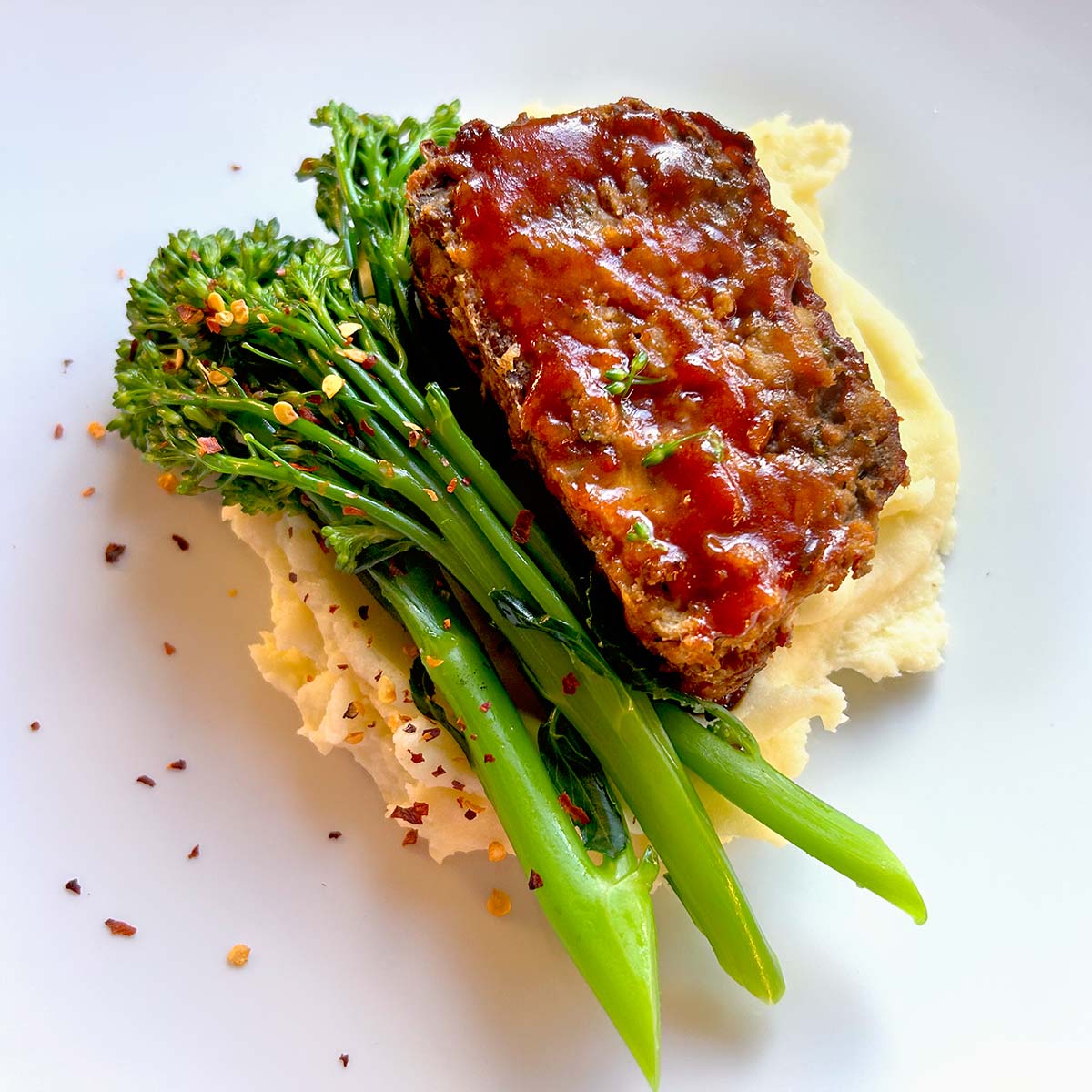 A slice of glazed meatloaf sits on creamy mashed potatoes, garnished with steamed tenderstem broccoli and sprinkled with red chilli flakes, served on a white plate.