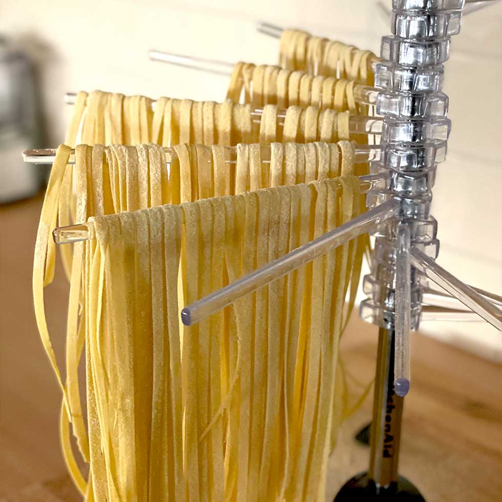 Freshly made pasta noodles hanging to dry on a clear plastic drying rack in a kitchen setting. The uncooked pasta strands are neatly arranged in rows.