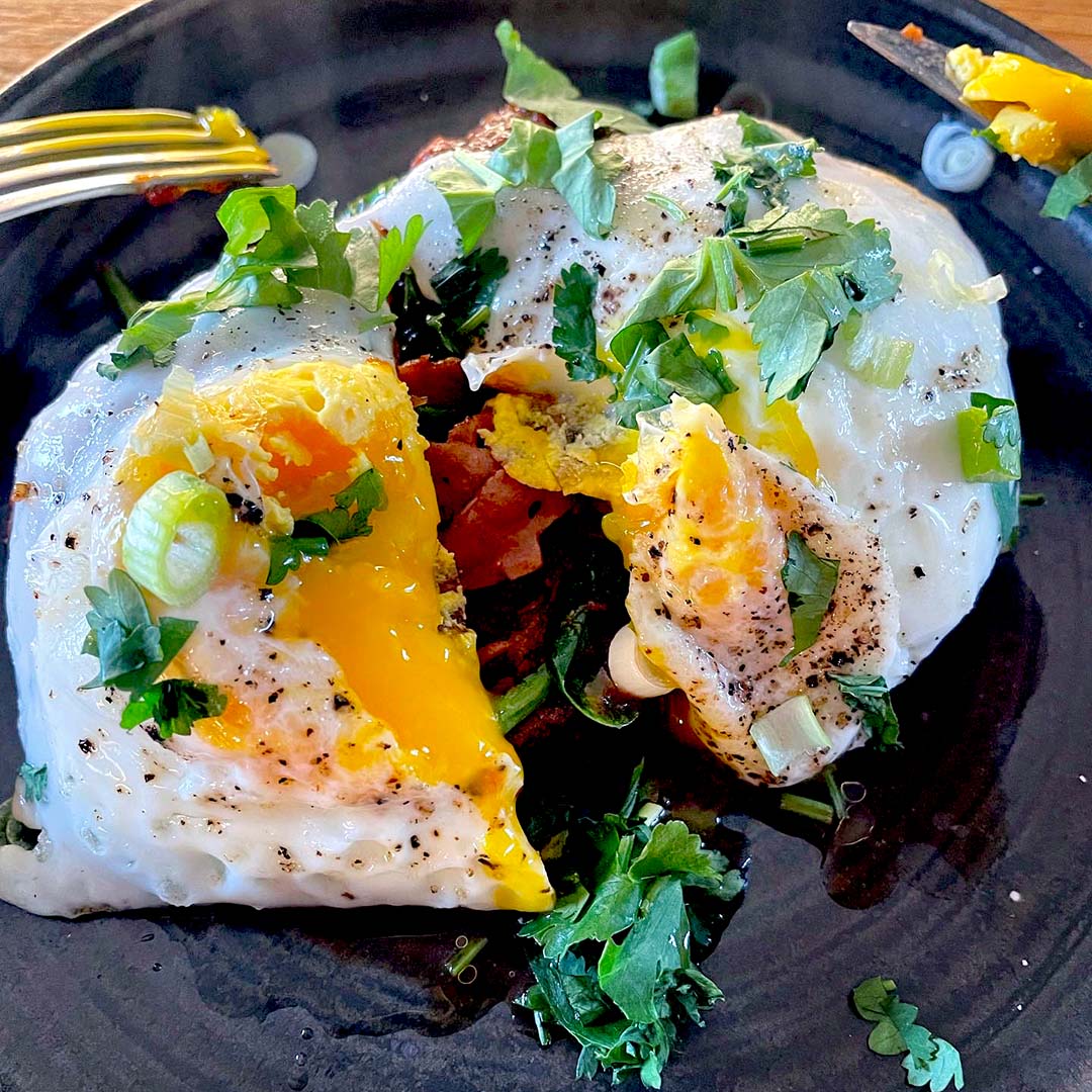Two fried eggs with runny yolks are garnished with fresh herbs and chopped spring onions, served on a dark plate. A fork cuts into one egg, letting the yolk flow out over sautéed vegetables underneath.