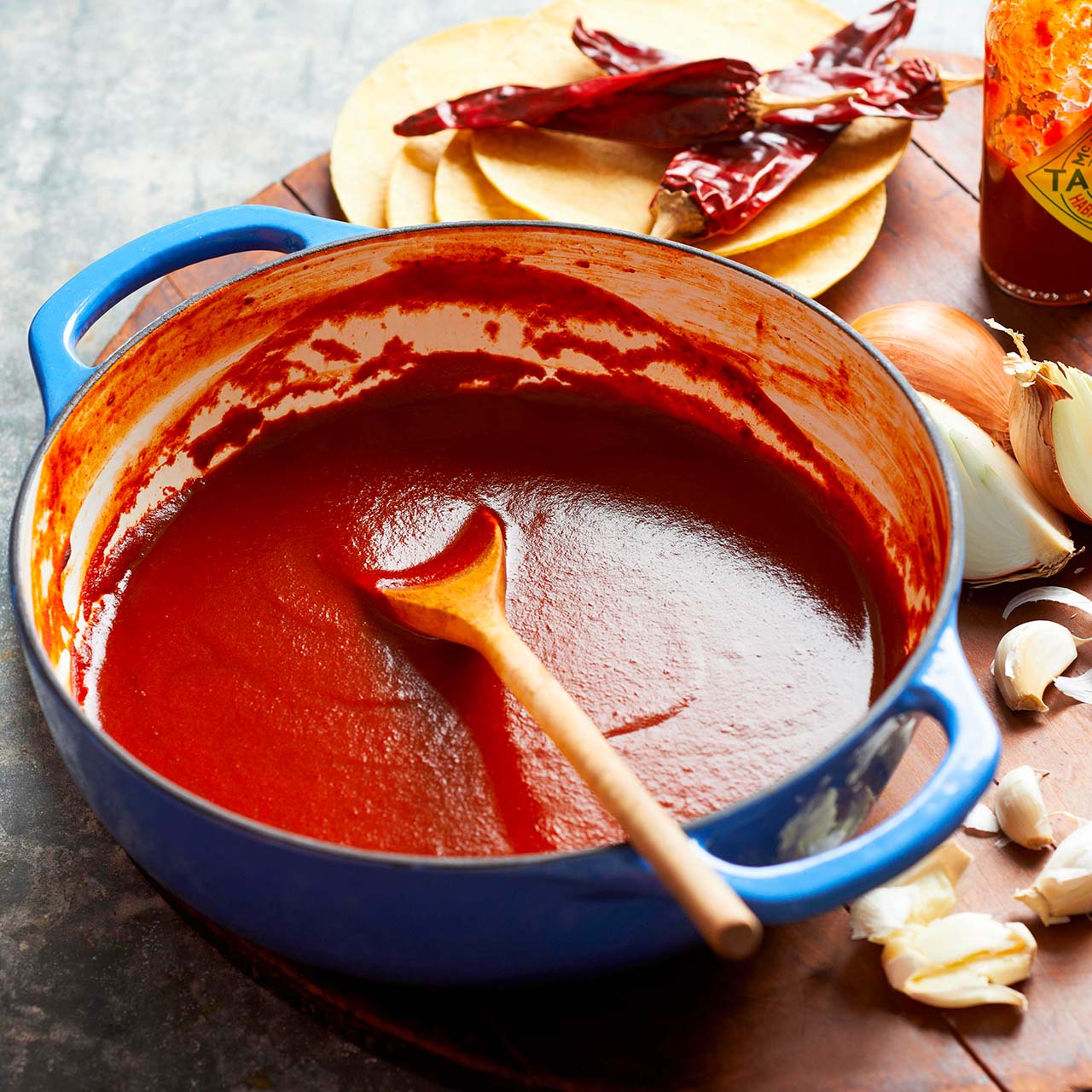 A blue pot filled with smooth red enchilada sauce sits on a wooden surface, surrounded by a wooden spoon, dried chillies, corn tortillas, a halved onion, garlic cloves, and a jar of hot sauce.