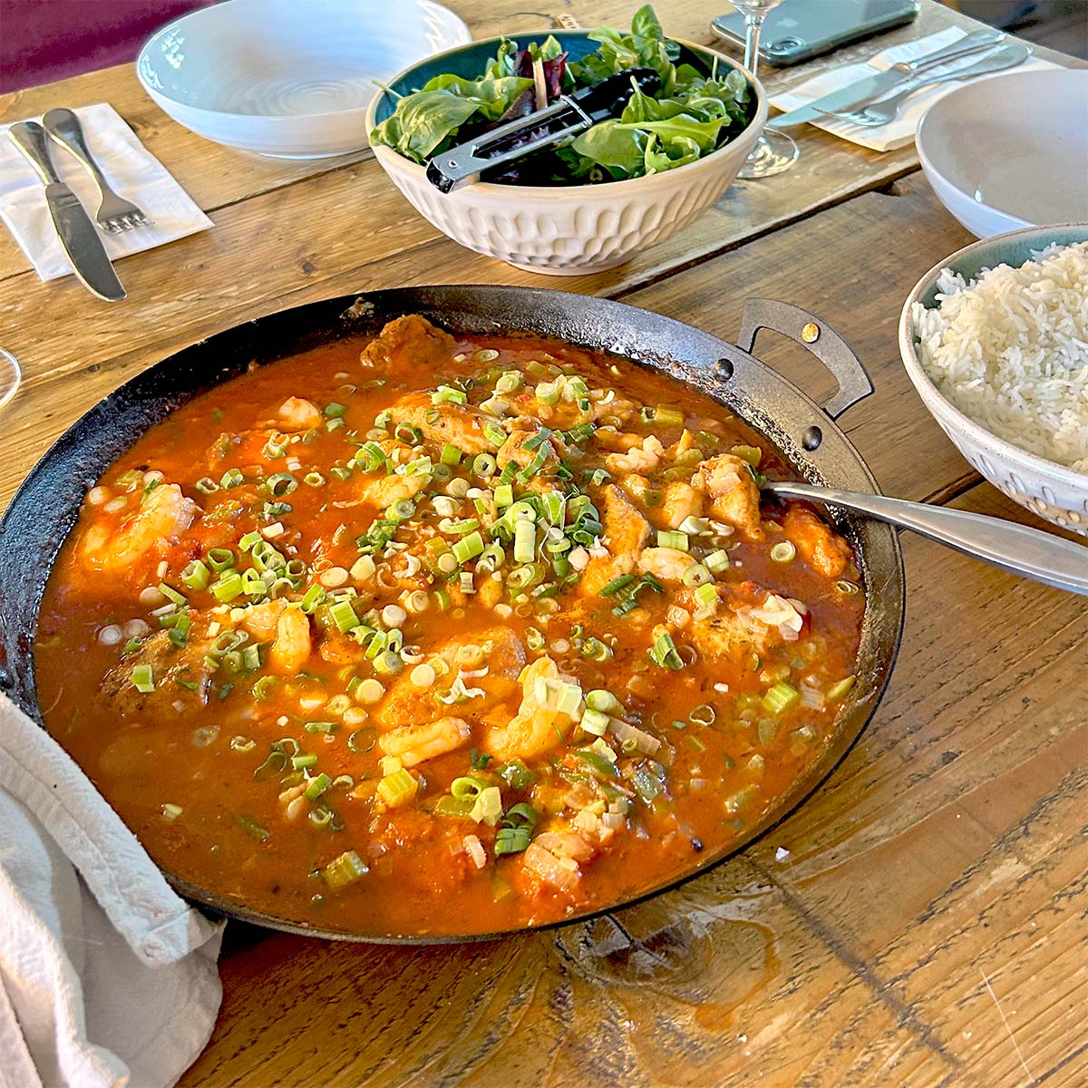 A frying pan filled with a red tomato-based stew topped with chopped spring onions sits on a wooden table, alongside a bowl of white rice, a salad, and empty plates with cutlery.