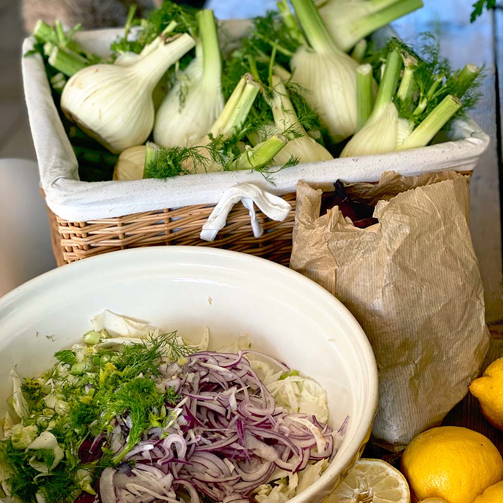 A wicker basket filled with fresh fennel bulbs sits behind a bowl containing sliced fennel, red onion, and herbs. A brown paper bag and two lemons are also on the table.
