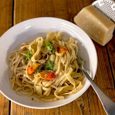 A white bowl of fettuccine pasta with cherry tomatoes, fresh herbs, and black pepper. A fork rests on the bowl, and a block of cheese with a grater is in the background on a wooden table.