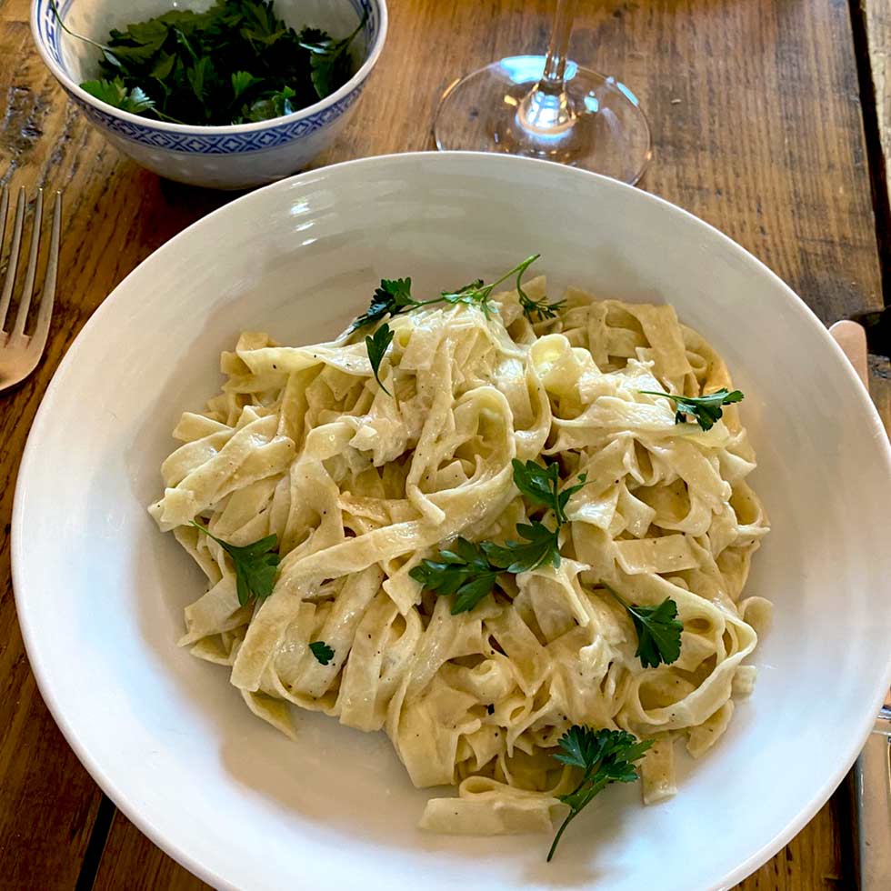 A bowl of fettuccine Alfredo topped with fresh parsley sits on a wooden table next to a bowl of chopped herbs, a glass of wine, and a fork.