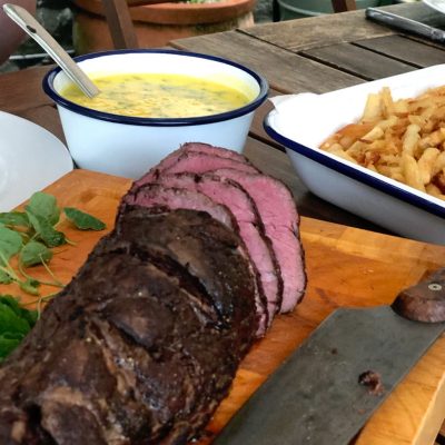 Sliced roast beef on a wooden chopping board with a knife, next to a bowl of chips and a bowl of yellow sauce with herbs, all set on an outdoor wooden table.