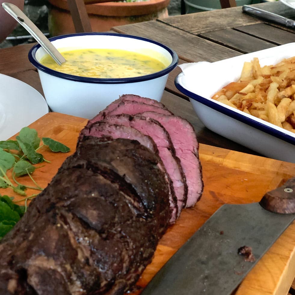 Sliced roast beef on a wooden chopping board with a knife, next to a bowl of chips and a bowl of yellow sauce with herbs, all set on an outdoor wooden table.