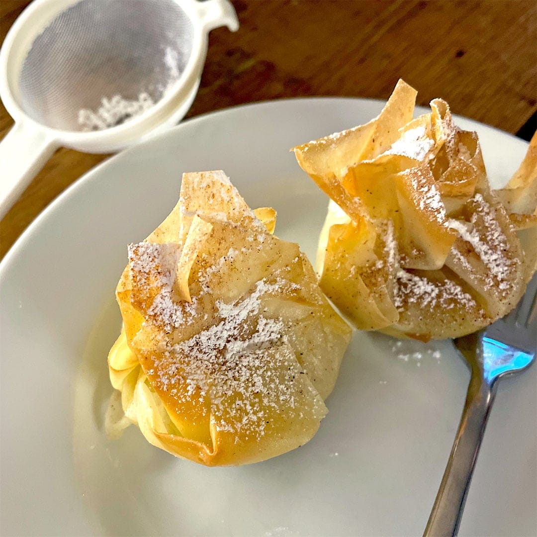 Two golden-brown pastry parcels dusted with icing sugar sit on a white plate with a fork; a mesh sieve with sugar is in the background on a wooden table.