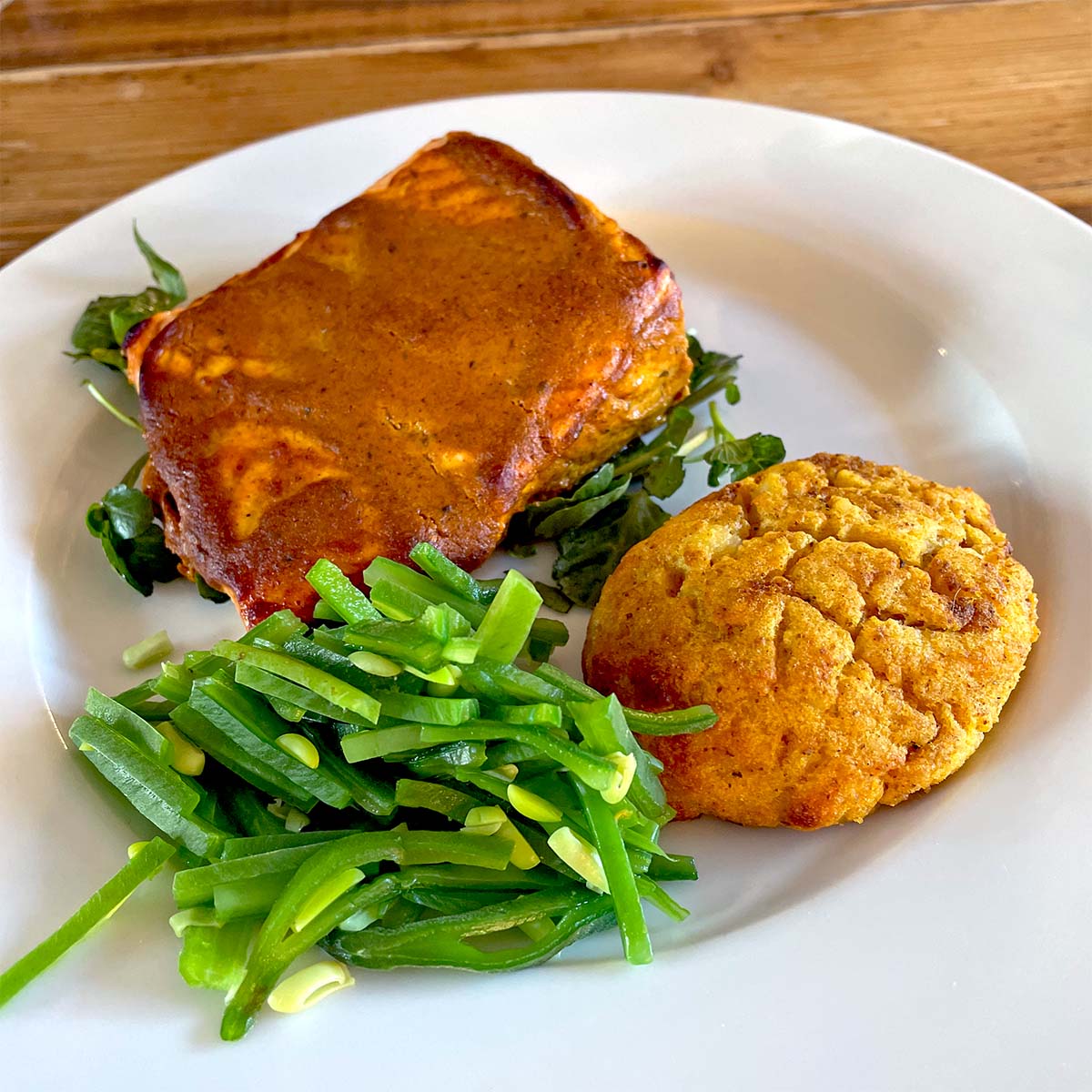 A white plate with a serving of lasagne on leafy greens, a scoop of sautéed green beans, and a golden-brown scone. The food is arranged neatly on a wooden table.