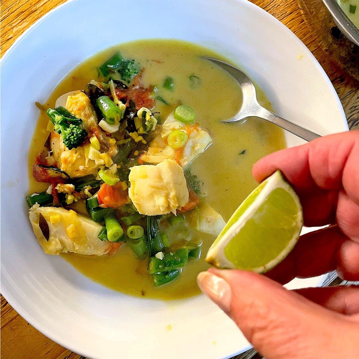 A hand squeezes a lime wedge over a bowl of fish and vegetable stew in a yellow broth, with a spoon resting inside the white bowl.
