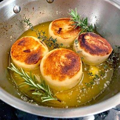 Four golden-brown, round potato slices are seared in a pan with melted butter, garnished with sprigs of fresh rosemary and thyme.