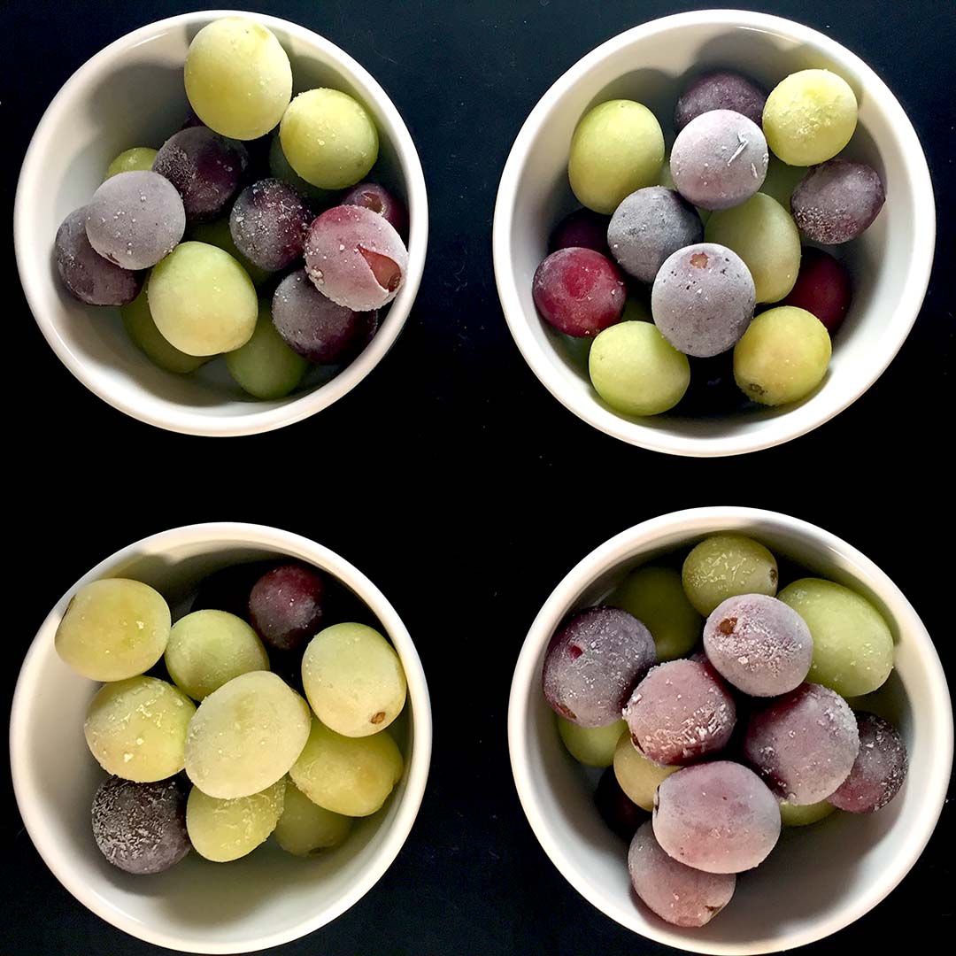 Four white bowls filled with green and purple grapes, some with a frosty appearance, arranged in a square pattern on a black background.