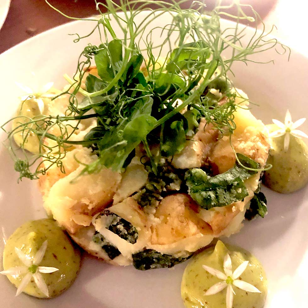 A plated dish featuring a creamy vegetable medley garnished with fresh herbs and edible flowers, accompanied by three dollops of green sauce, each topped with a small white flower.
