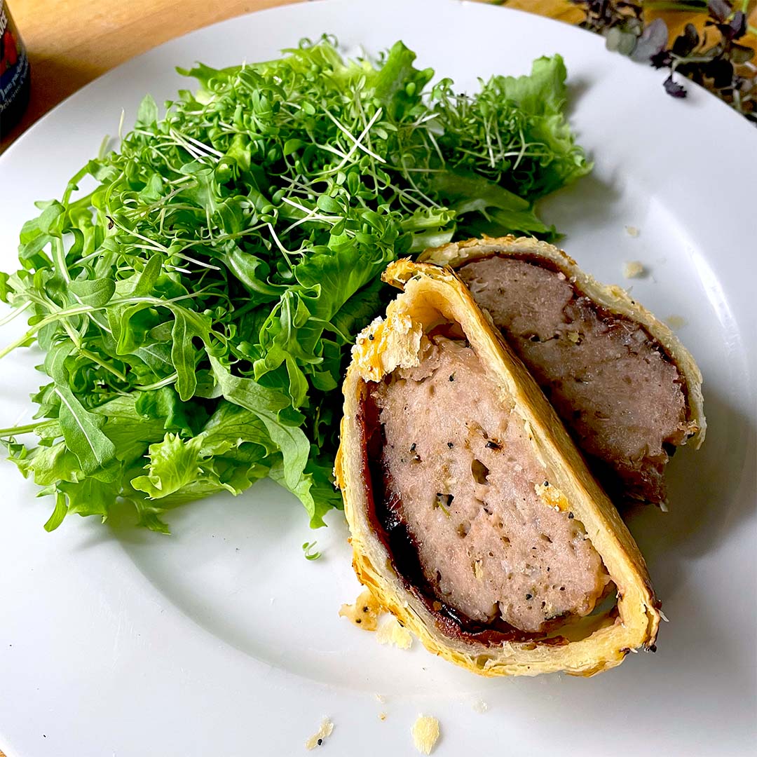 A plate with a leafy green salad and a sliced portion of meat wrapped in golden puff pastry, resembling a homemade pork Wellington, on a white plate.