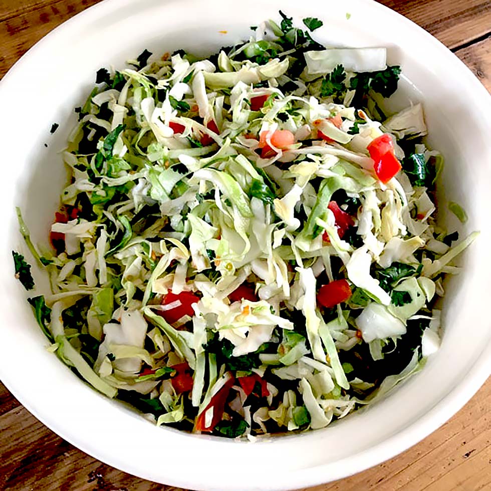 A white bowl filled with shredded cabbage salad mixed with chopped tomatoes, coriander, and green herbs, placed on a wooden surface.