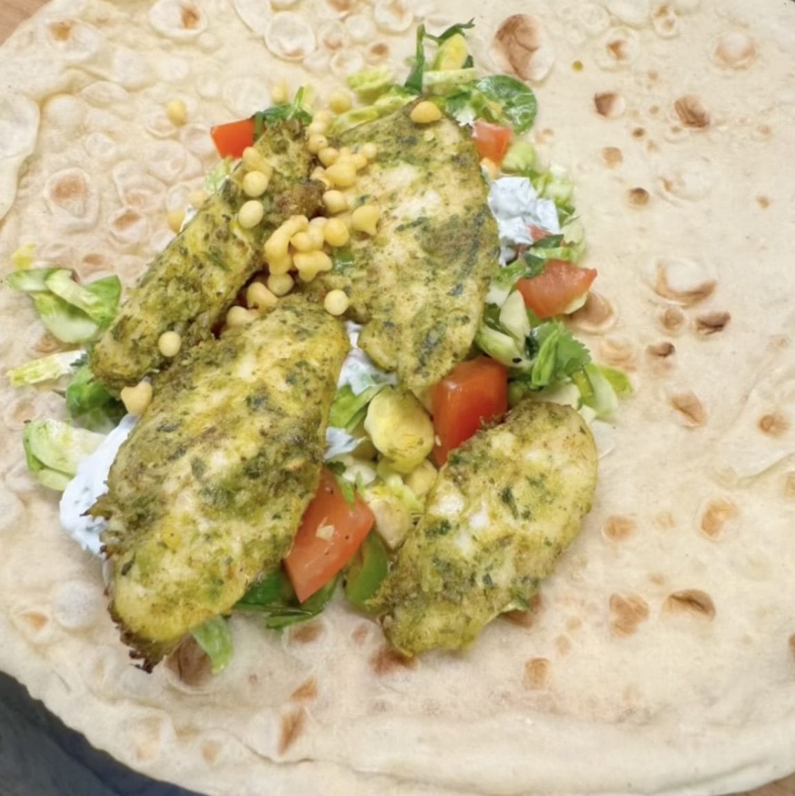 An open flatbread wrap topped with herbed chicken pieces, chopped lettuce, diced tomatoes, sweetcorn kernels, and a white sauce, ready to be folded.