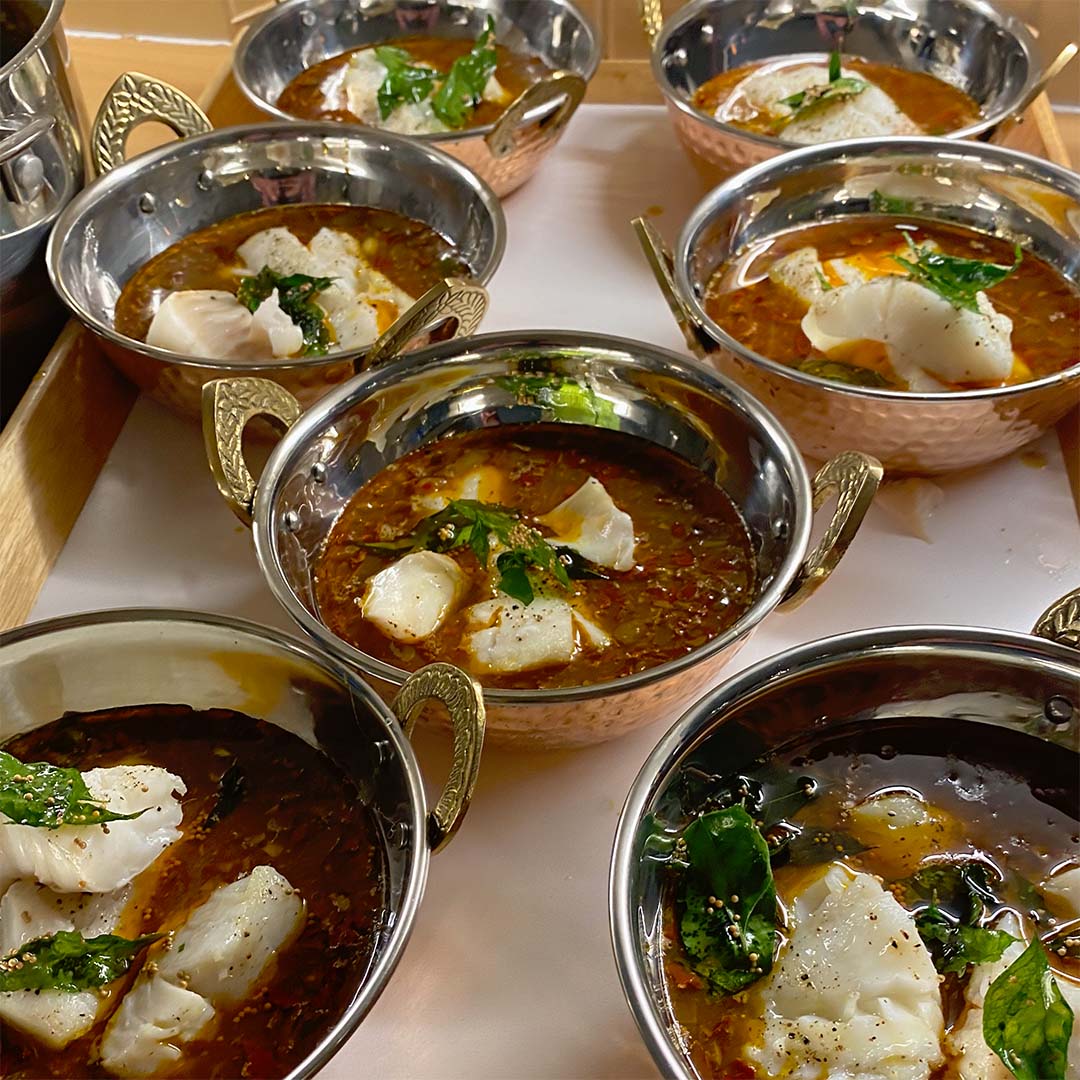 Six metal bowls filled with spicy red curry, garnished with herbs and chunks of white fish, are arranged on a tray. The curry has a rich, oily surface and each bowl has ornate metal handles.