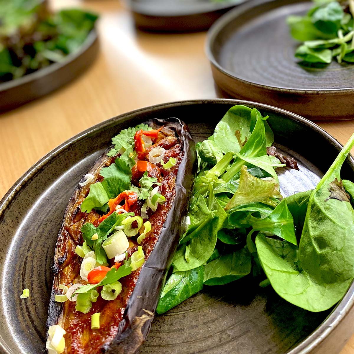 A dark plate with a roasted aubergine half topped with chopped spring onions, red chilli, and herbs, served alongside a fresh green salad.