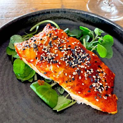 A fillet of salmon topped with a red glaze and garnished with white and black sesame seeds, served on a bed of fresh green leaves on a dark plate.