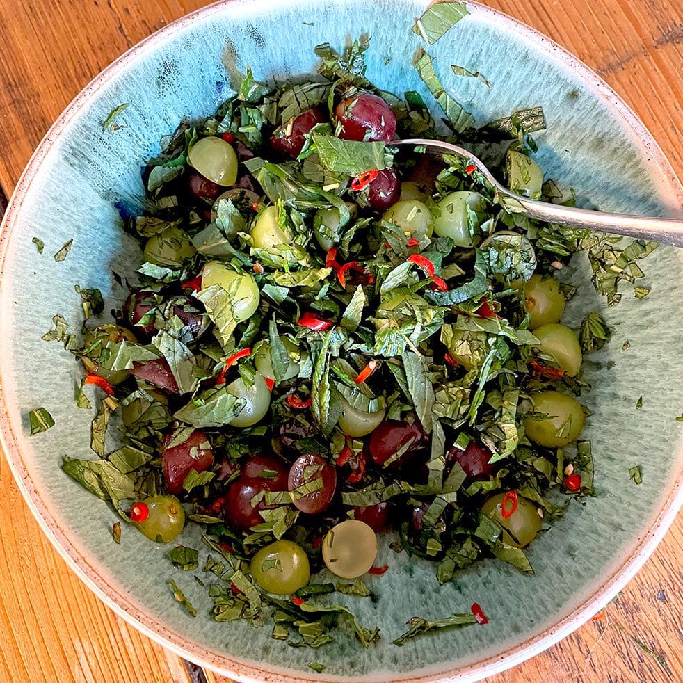 A bowl containing chopped green herbs, red and green grapes, sliced red chilli, and black olives, with a spoon resting in the mixture on a wooden surface.