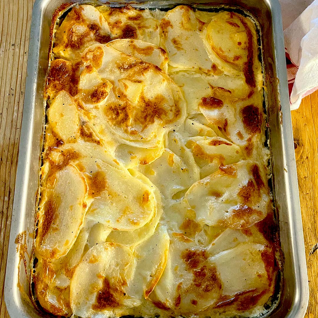 A baked potato gratin in a rectangular metal dish, featuring thinly sliced potatoes layered with cream and cheese, with a golden-brown, slightly crispy top. The dish sits on a wooden surface.