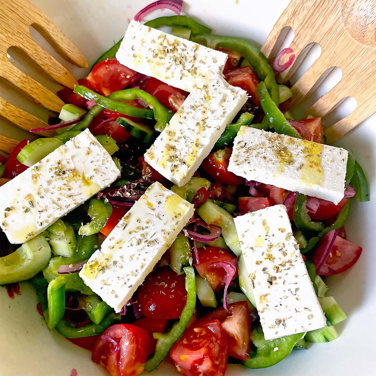 A bowl of Greek salad with sliced tomatoes, cucumbers, green peppers, red onions, and five rectangular pieces of feta cheese, garnished with dried herbs and olive oil. Wooden salad servers rest at the side.