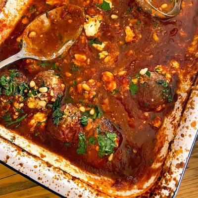 A baking dish filled with meatballs in a rich tomato sauce, topped with crumbled cheese, chopped herbs, and pine nuts. Two metal serving spoons rest in the dish.