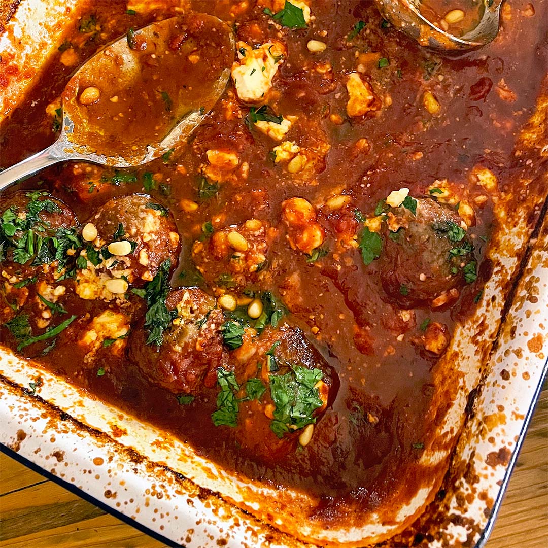 A baking dish filled with meatballs in a rich tomato sauce, topped with crumbled cheese, chopped herbs, and pine nuts. Two metal serving spoons rest in the dish.